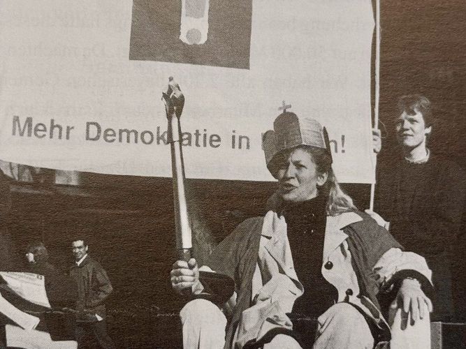Aktion Der Bürger wird König/Die Bürgerin wird Königin während des Volkksbegeherens Mehr Demokratie in Bayern 1995. Man sieht eine zur Königin gekrönte Bürgerin mit einem Szepter, auf einem Thron sitzend. Im  Hintergrund ein Plakat mit der Aufschrift Mehr Demokratie in Bayern und dem Logo mit dem Ausrufezeichen.